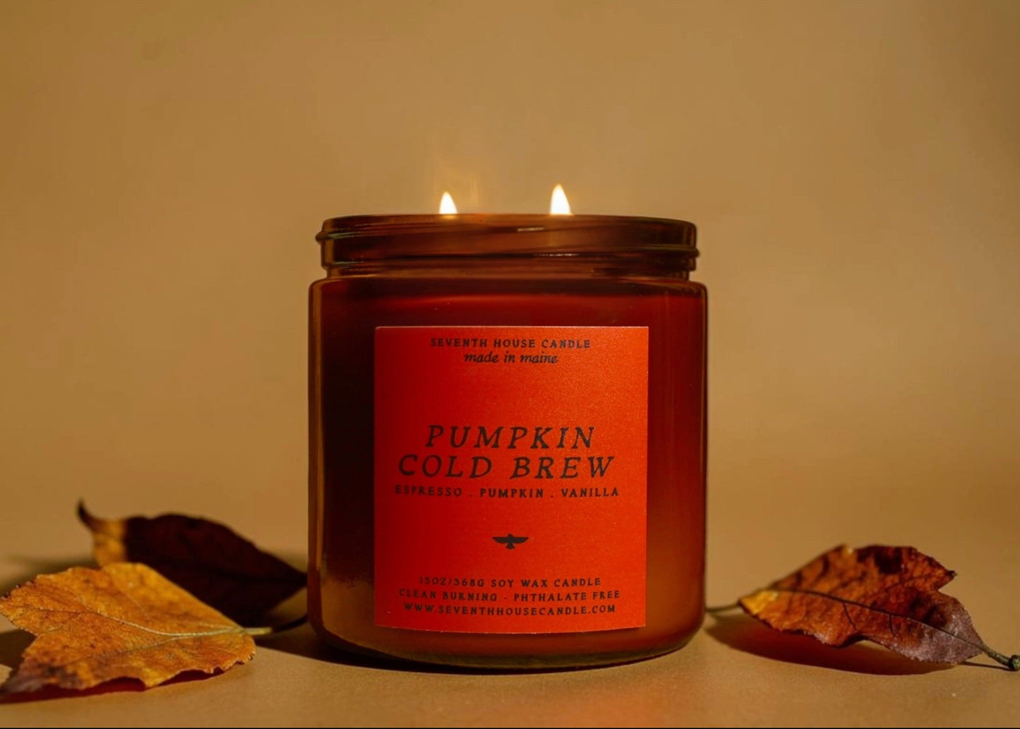 A lit double-wick candle in a dark amber glass jar with a warm orange label reading 'Pumpkin Cold Brew' and 'Desert Rose Candle.' The candle is photographed on a beige surface against a beige background, with dried autumn leaves placed near the base.