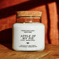 A white soy wax candle in a clear glass jar with a cork lid, labeled 'Apple of My Pie' by Seventh House Candle, set on a wooden surface with warm, dramatic sunlight and shadows.