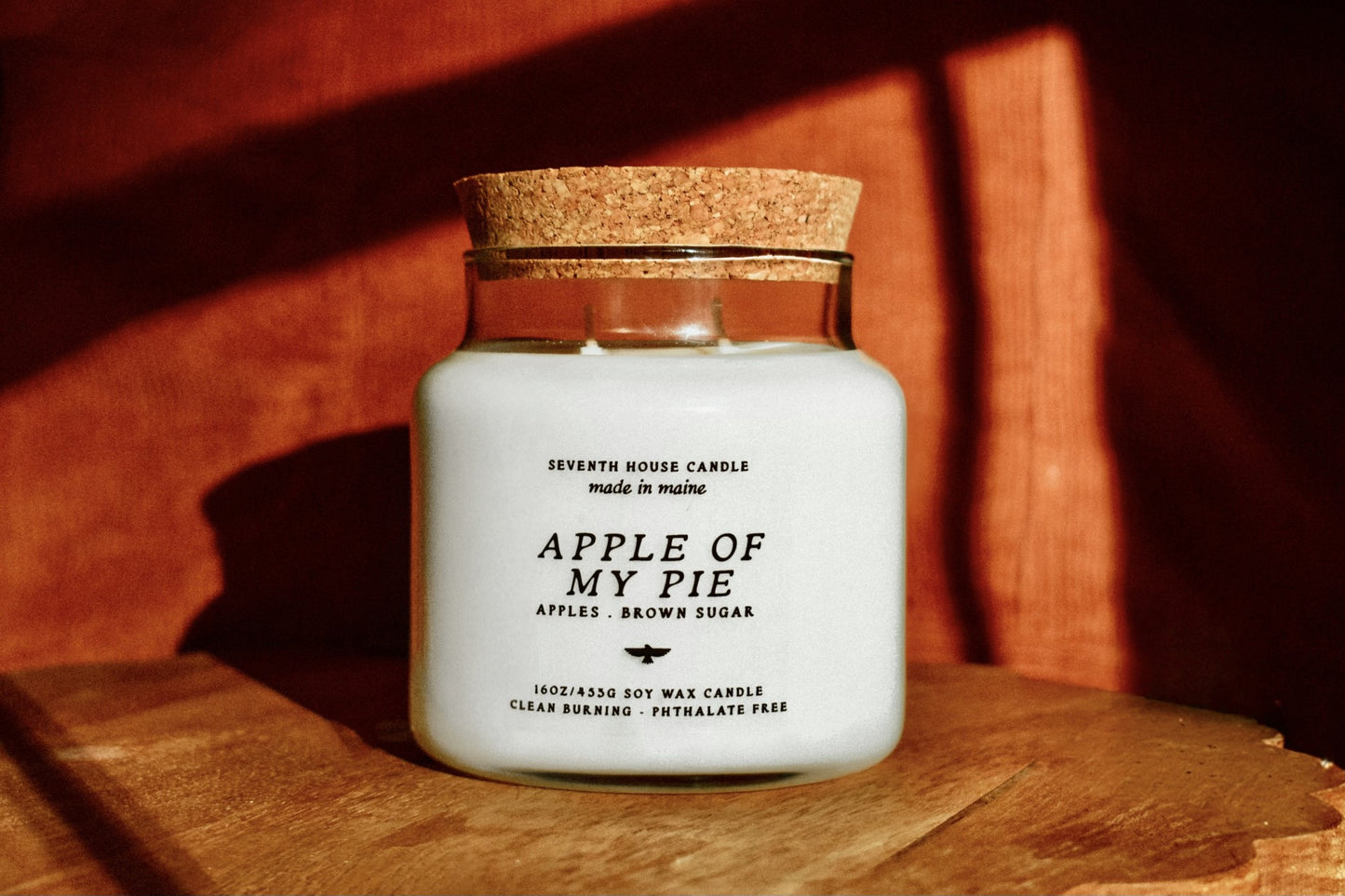 A white soy wax candle in a clear glass jar with a cork lid, labeled 'Apple of My Pie' by Seventh House Candle, set on a wooden surface with warm, dramatic sunlight and shadows.