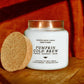 A lit white candle in a clear glass jar with a cork lid leaning against it on a wooden surface, set against a warm orange-brown draped fabric background. The label reads 'Pumpkin Cold Brew' by Seventh House Candle.