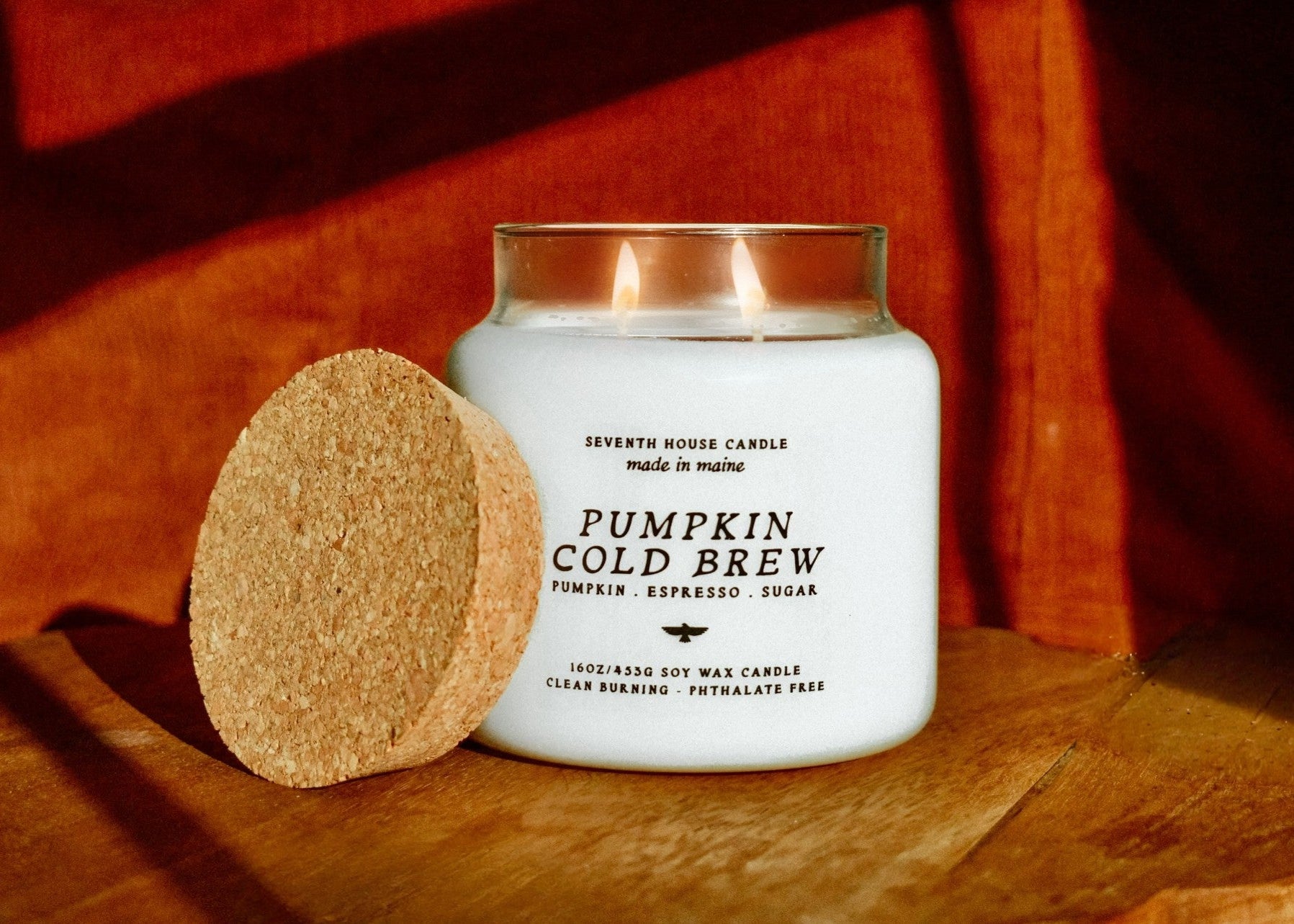 A lit white candle in a clear glass jar with a cork lid leaning against it on a wooden surface, set against a warm orange-brown draped fabric background. The label reads 'Pumpkin Cold Brew' by Seventh House Candle.