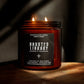 A lit amber glass jar candle with two flames, featuring a black label that reads 'Haunted Library Leather & Teakwood' by Seventh House Candle, set on a wooden surface against a dark background with dramatic shadows.