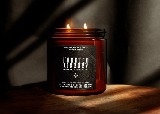 A lit amber glass jar candle with two flames, featuring a black label that reads 'Haunted Library Leather & Teakwood' by Seventh House Candle, set on a wooden surface against a dark background with dramatic shadows.