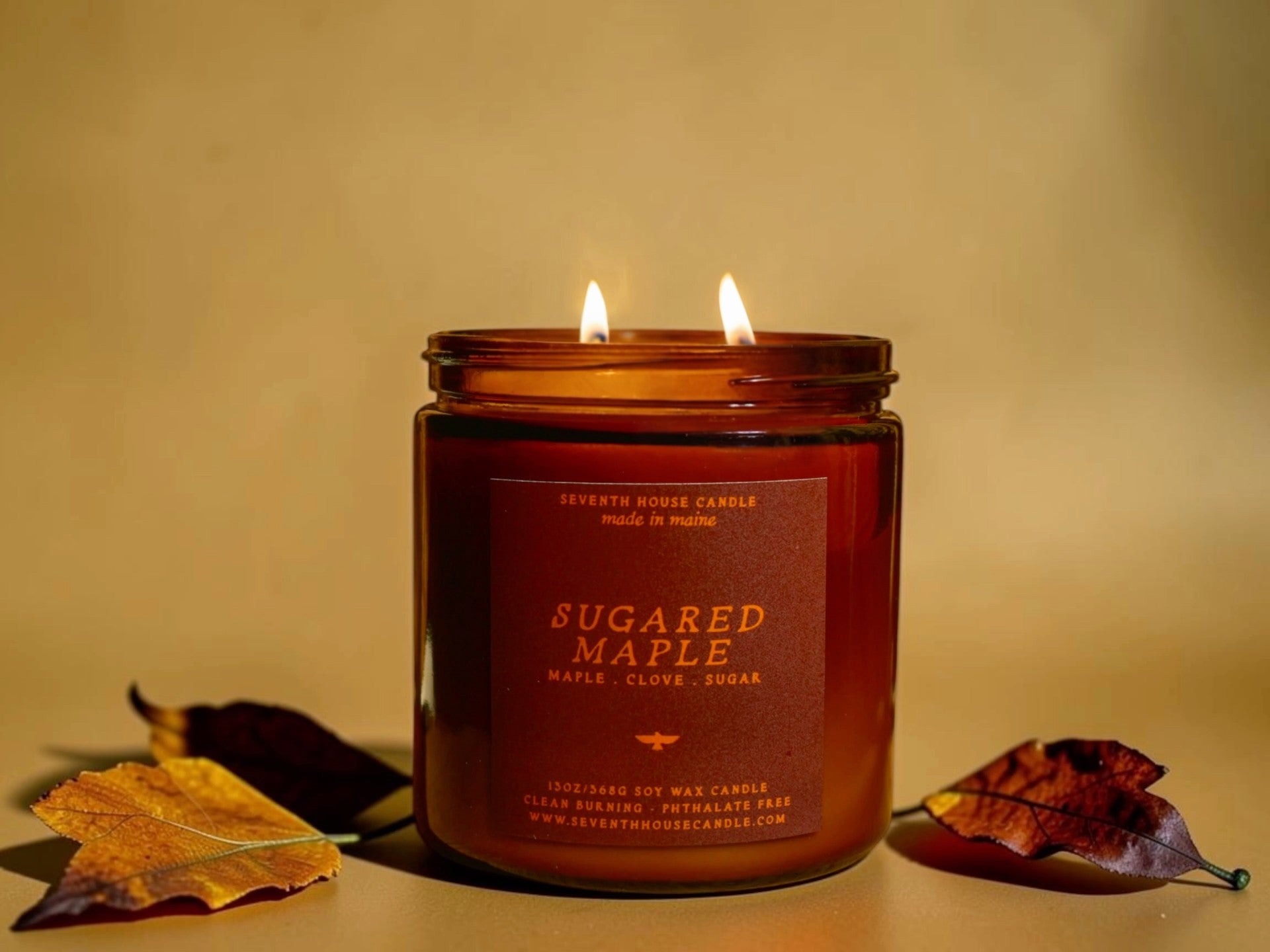 A lit amber glass jar candle labeled 'SUGARED MAPLE' with two flames burning, set against a warm beige background with decorative autumn leaves on either side.