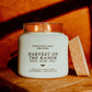 A lit white candle in a clear glass jar with the label 'Harvest on the Ranch' by Seventh House Candle, placed on a wooden surface with warm sunlight and shadows. A textured brown block stands beside the jar.