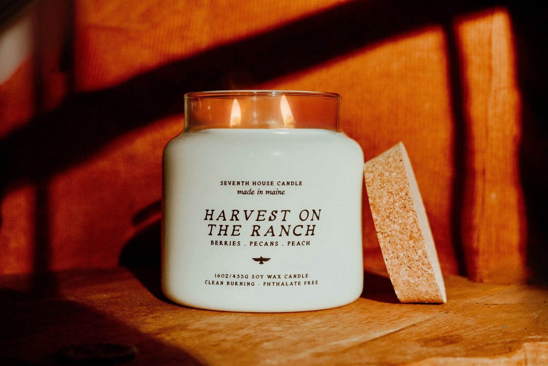 A lit white candle in a clear glass jar with the label 'Harvest on the Ranch' by Seventh House Candle, placed on a wooden surface with warm sunlight and shadows. A textured brown block stands beside the jar.