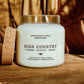 A white soy wax candle in a clear glass jar with two lit wicks, labeled 'HIGH COUNTRY' by Seventh House Candle, placed on a wooden surface with a cork coaster beside it and a warm brown fabric background.