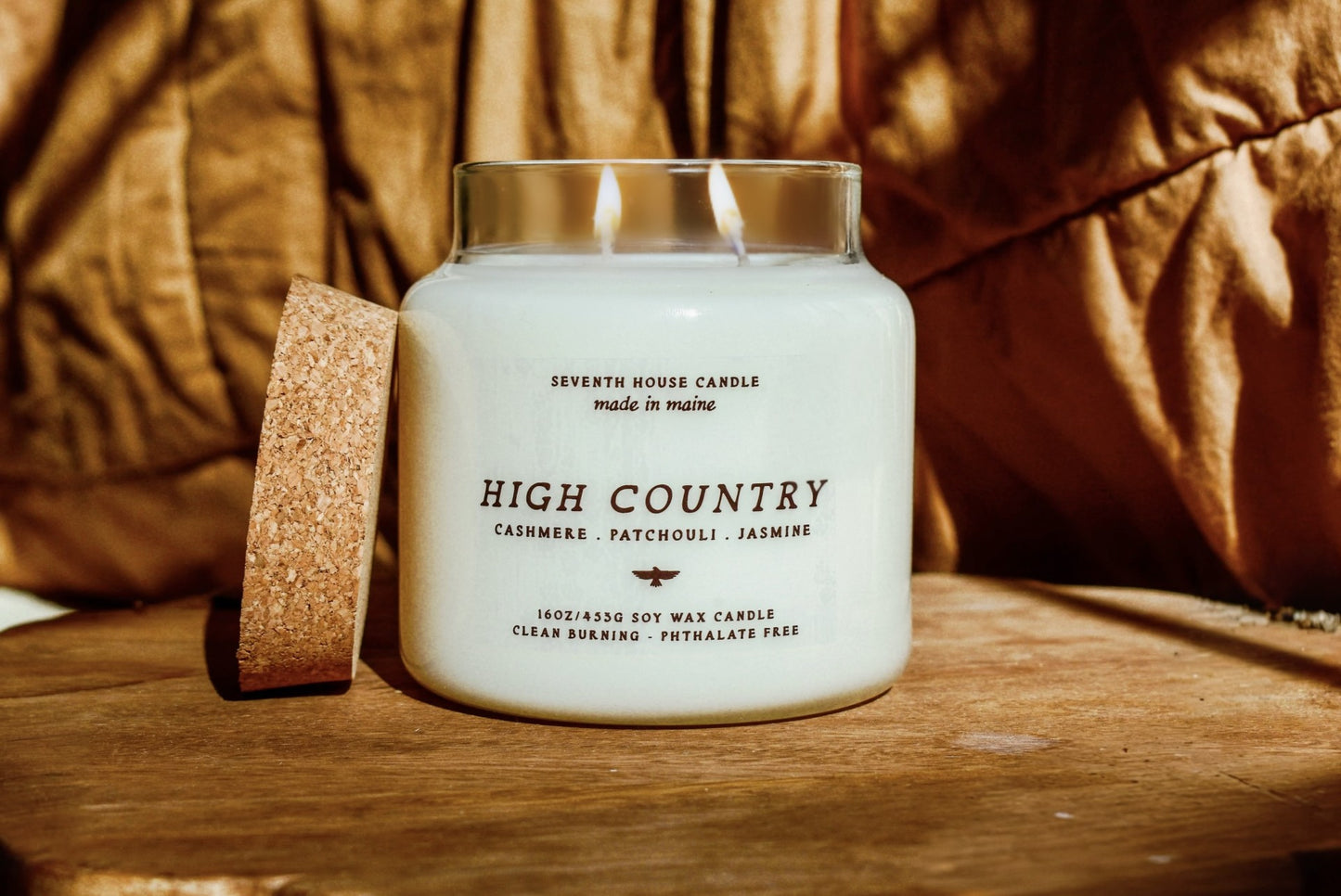 A white soy wax candle in a clear glass jar with two lit wicks, labeled 'HIGH COUNTRY' by Seventh House Candle, placed on a wooden surface with a cork coaster beside it and a warm brown fabric background.