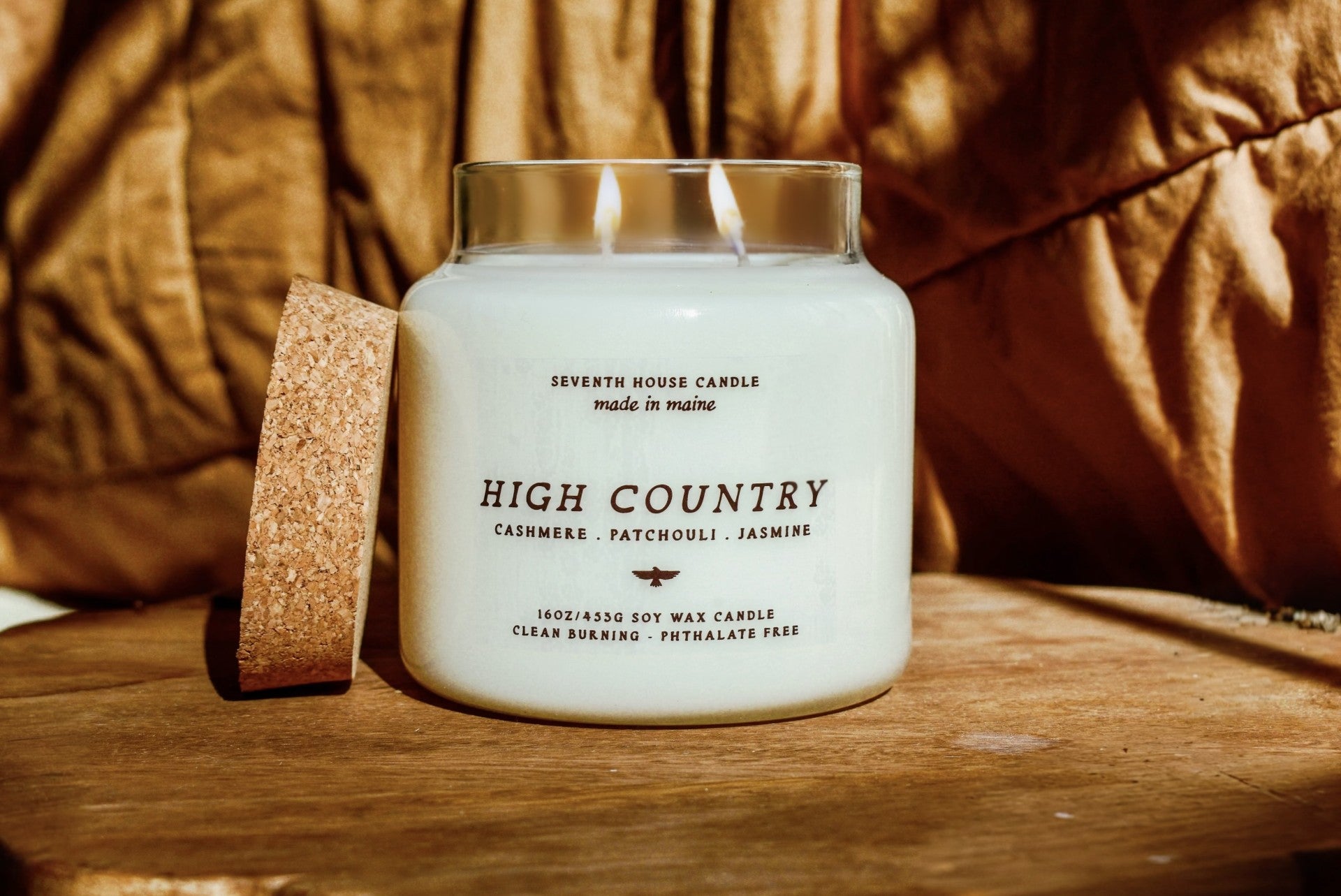 A white soy wax candle in a clear glass jar with two lit wicks, labeled 'HIGH COUNTRY' by Seventh House Candle, placed on a wooden surface with a cork coaster beside it and a warm brown fabric background.