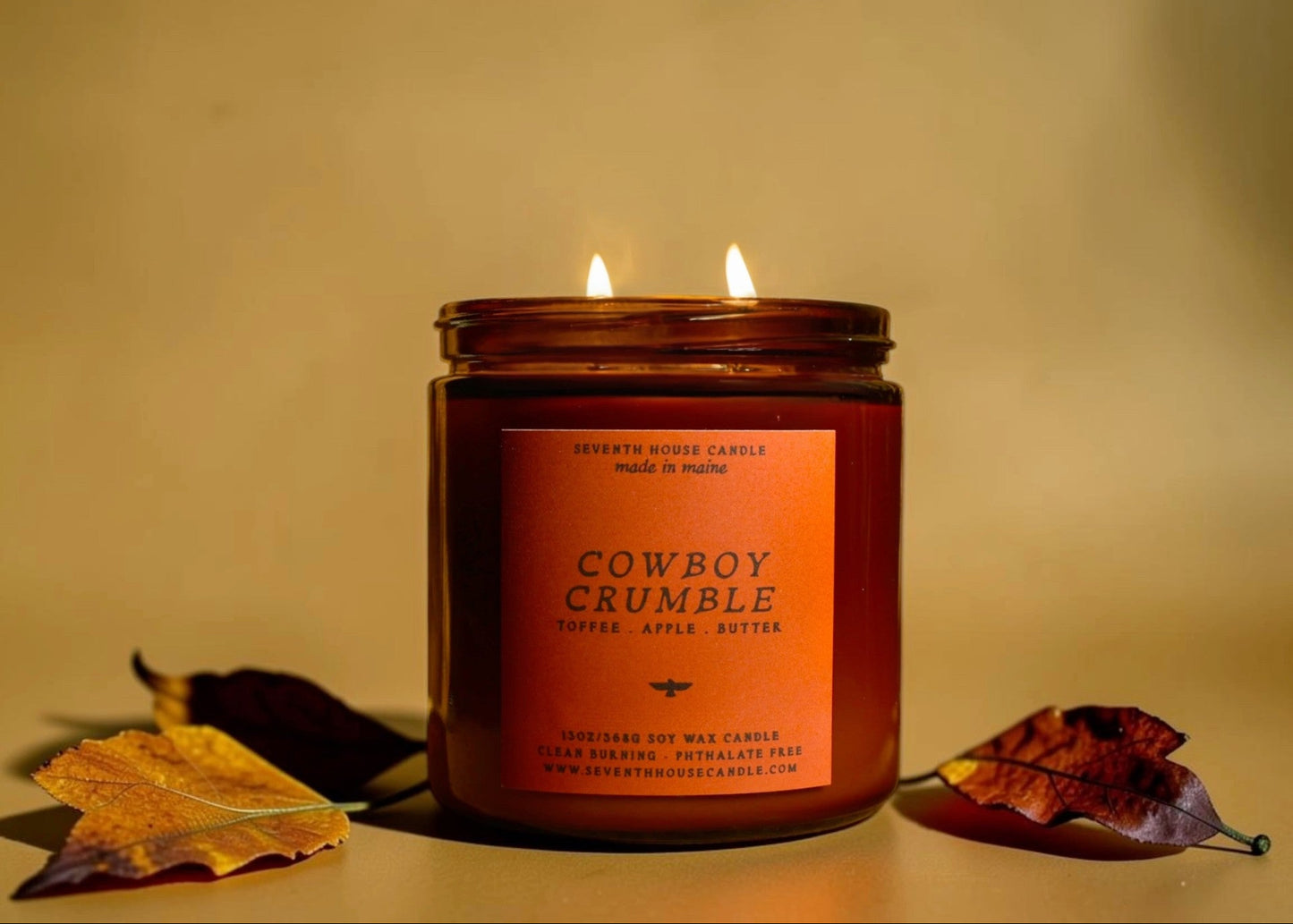 A lit amber glass jar candle with two flames, labeled 'Cowboy Crumble' by Seventh House Candle, styled on a beige surface with autumn leaves against a warm beige background.