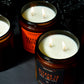 Four lit amber glass jar candles with white wax and black labels, arranged on a dark surface against a black background. Visible label text includes 'Wicked in the West,' 'Bourbon Barrel,' and 'Twilight Torch.