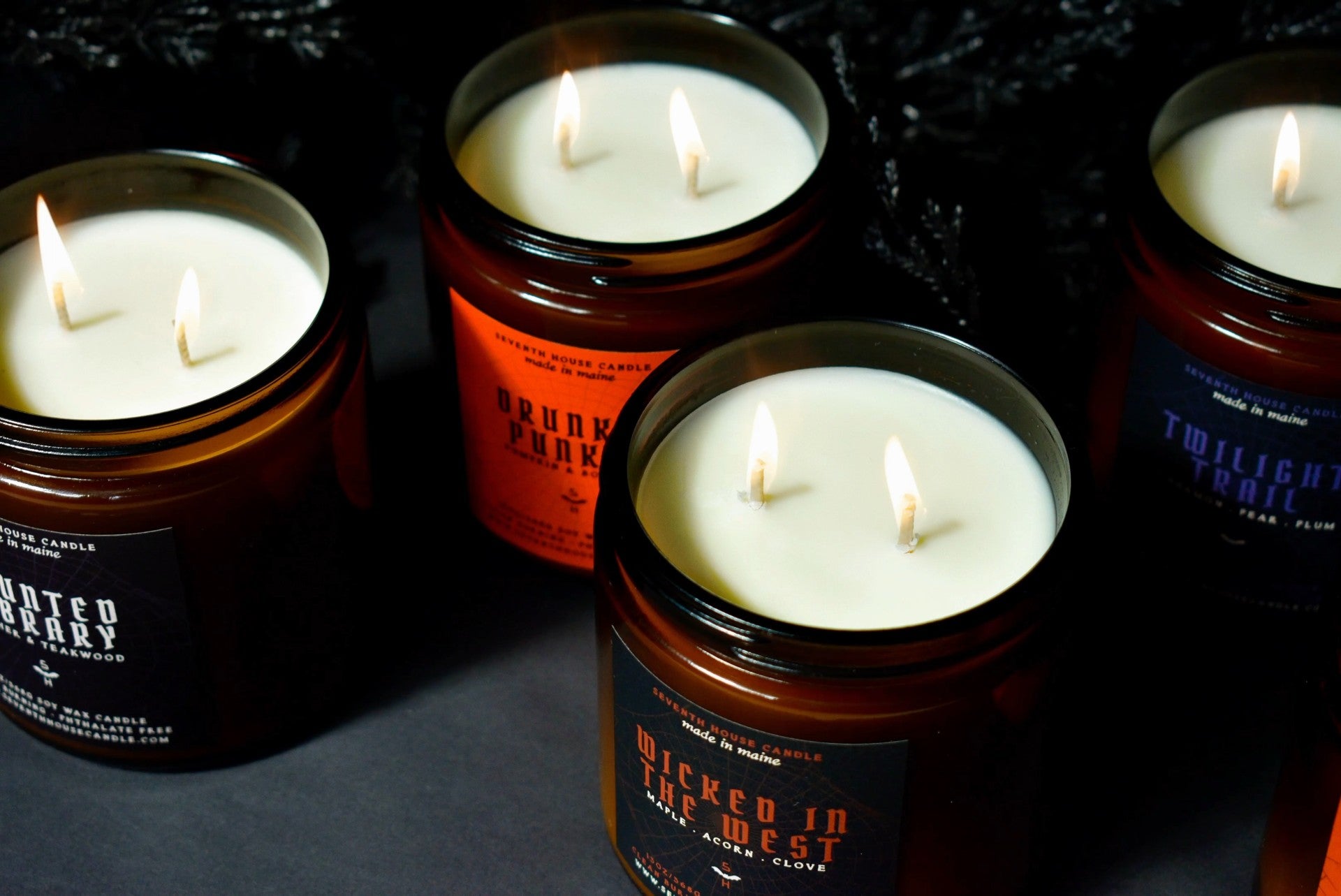 Four lit amber glass jar candles with white wax and black labels, arranged on a dark surface against a black background. Visible label text includes 'Wicked in the West,' 'Bourbon Barrel,' and 'Twilight Torch.
