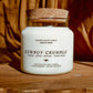 A white soy wax candle in a clear glass jar with a cork stopper, labeled 'COWBOY CRUMBLE' by Seventh House Candle, set on a rustic wooden surface with a draped brown fabric background.