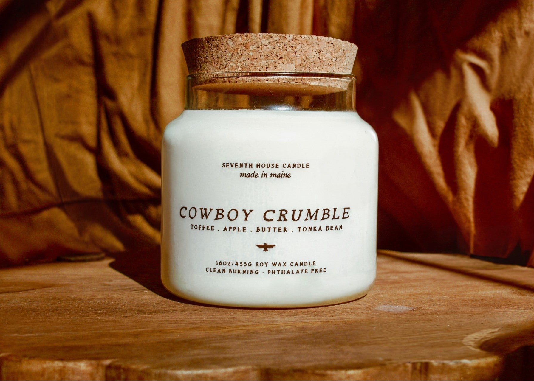 A white soy wax candle in a clear glass jar with a cork stopper, labeled 'COWBOY CRUMBLE' by Seventh House Candle, set on a rustic wooden surface with a draped brown fabric background.