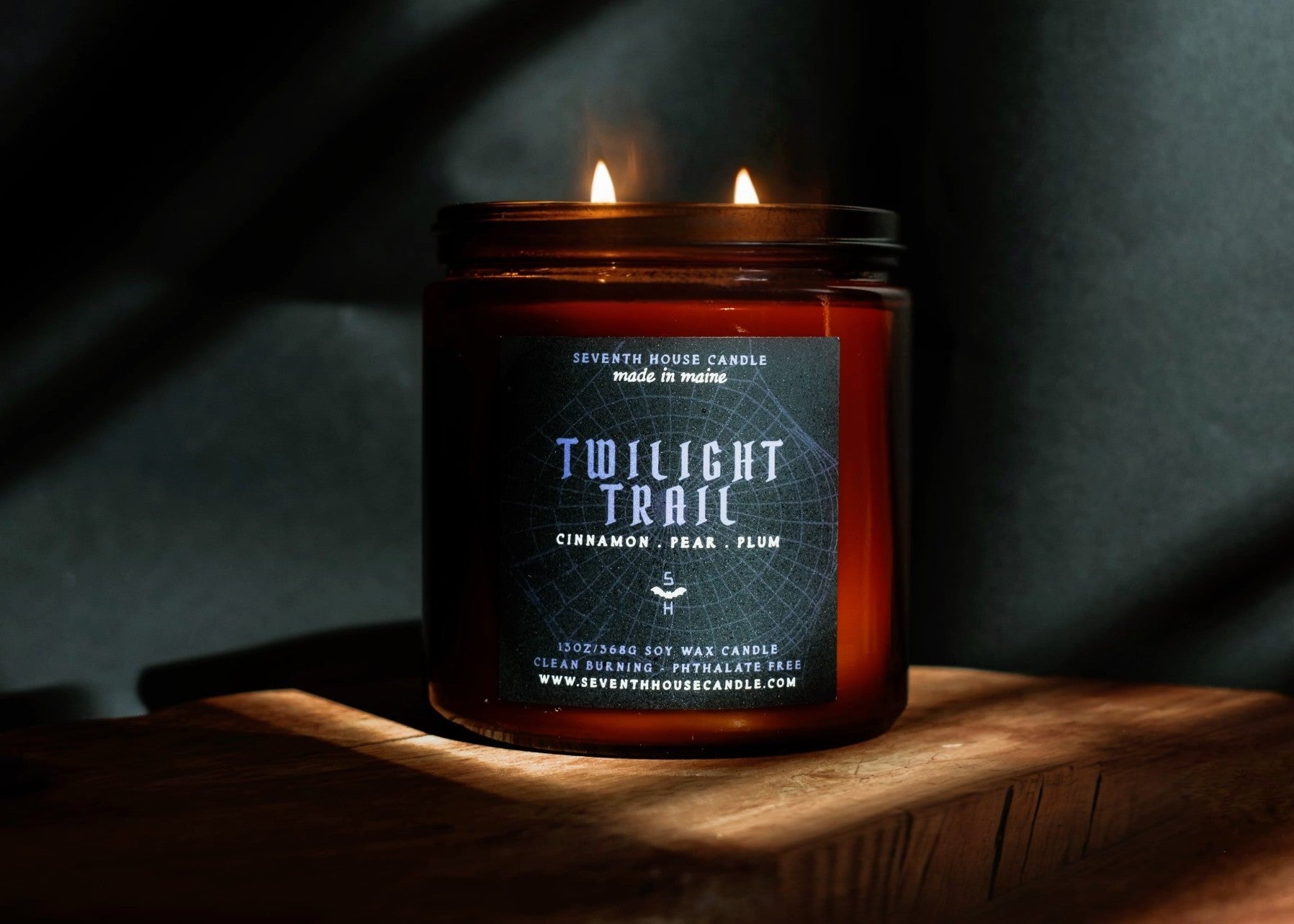 A lit amber glass jar candle labeled 'Twilight Trail' by Seventh House Candle, placed on a wooden surface against a dark draped background.