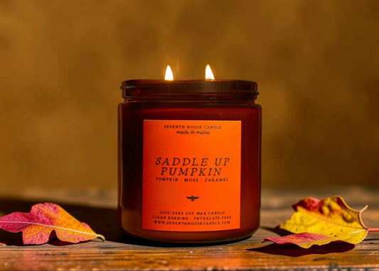 A lit amber glass jar candle labeled 'Saddle Up Pumpkin' sits on a rustic wooden surface with autumn leaves around it against a warm brown background.