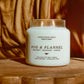 A lit white soy wax candle in a clear glass jar sits on a wooden surface against a warm brown draped fabric background. The label reads 'FOG & FLANNEL' with smaller text below including 'SEEDAMUT', 'MANJURY', and 'JASMINE'.