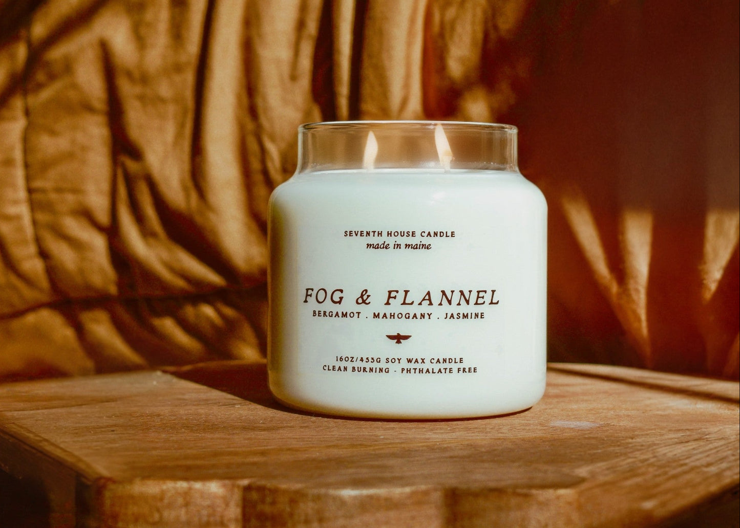 A lit white soy wax candle in a clear glass jar sits on a wooden surface against a warm brown draped fabric background. The label reads 'FOG & FLANNEL' with smaller text below including 'SEEDAMUT', 'MANJURY', and 'JASMINE'.