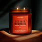 A lit two-wick soy wax candle in a dark amber glass jar with a terracotta-colored label reading 'October Mourning' and 'Cinnamon - Cassis Root,' set on a wooden surface against a dark background with dramatic shadows.
