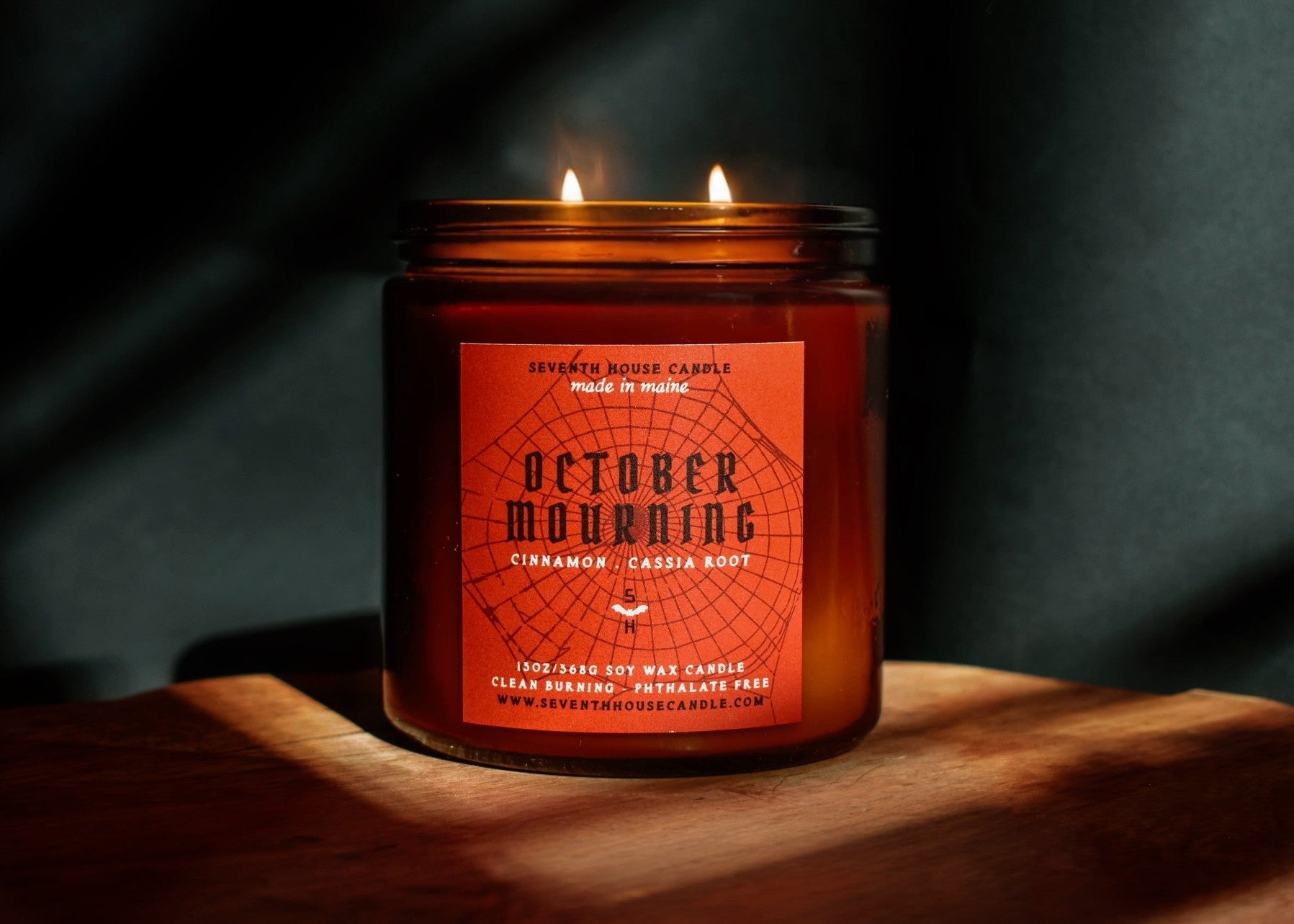 A lit two-wick soy wax candle in a dark amber glass jar with a terracotta-colored label reading 'October Mourning' and 'Cinnamon - Cassis Root,' set on a wooden surface against a dark background with dramatic shadows.