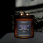A lit amber glass candle jar with a dark label reading 'Twilight Trail' and 'Cinnamon, Pear, Elm' scent, placed on a dark surface with moody shadows and dried foliage in the background.