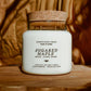 A white jar candle with a cork lid sits on a wooden surface against a warm, draped wood background. The label reads 'SUGARED MAPLE' and 'SEVENTH HOUSE CANDLE'.