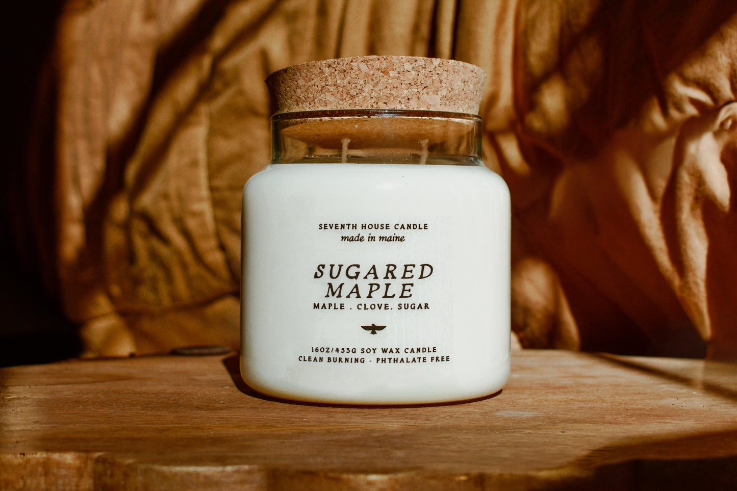 A white jar candle with a cork lid sits on a wooden surface against a warm, draped wood background. The label reads 'SUGARED MAPLE' and 'SEVENTH HOUSE CANDLE'.