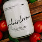 Candle labeled 'Heirloom' surrounded by tomatoes on a wooden surface