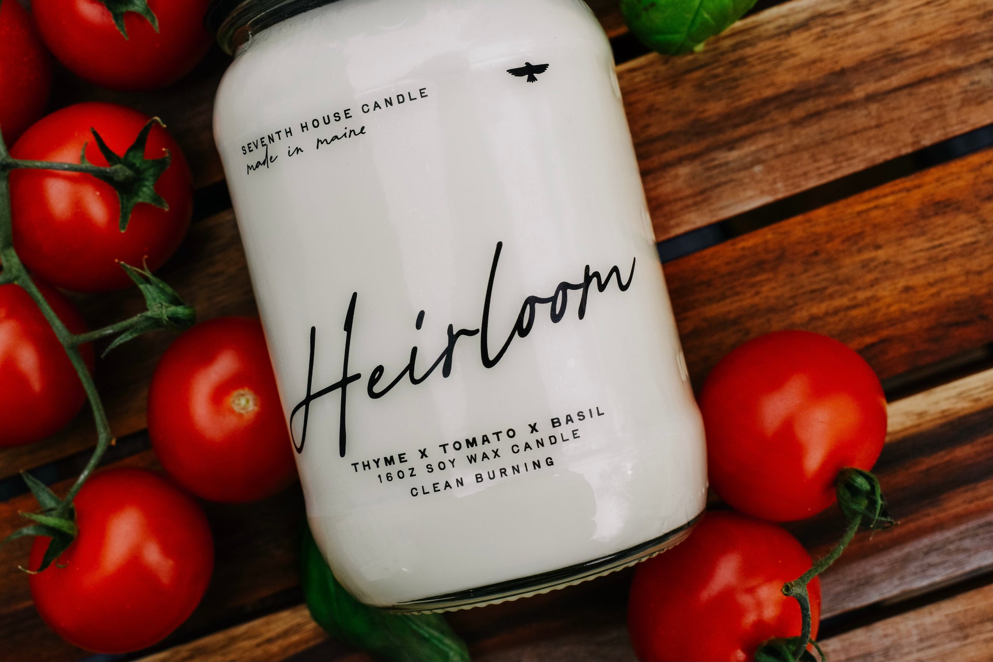 Candle labeled 'Heirloom' surrounded by tomatoes on a wooden surface