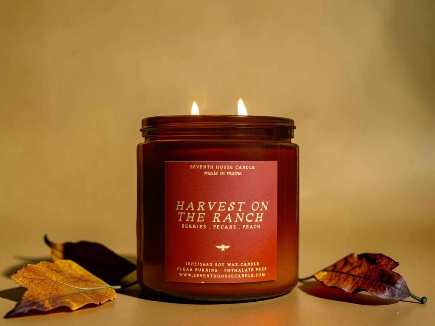 A lit double-wick soy wax candle in a dark amber glass jar with a red-orange label reading 'Harvest on the Ranch' and 'Seventh House Candle.' The candle is styled on a warm beige surface with autumn leaves on both sides.