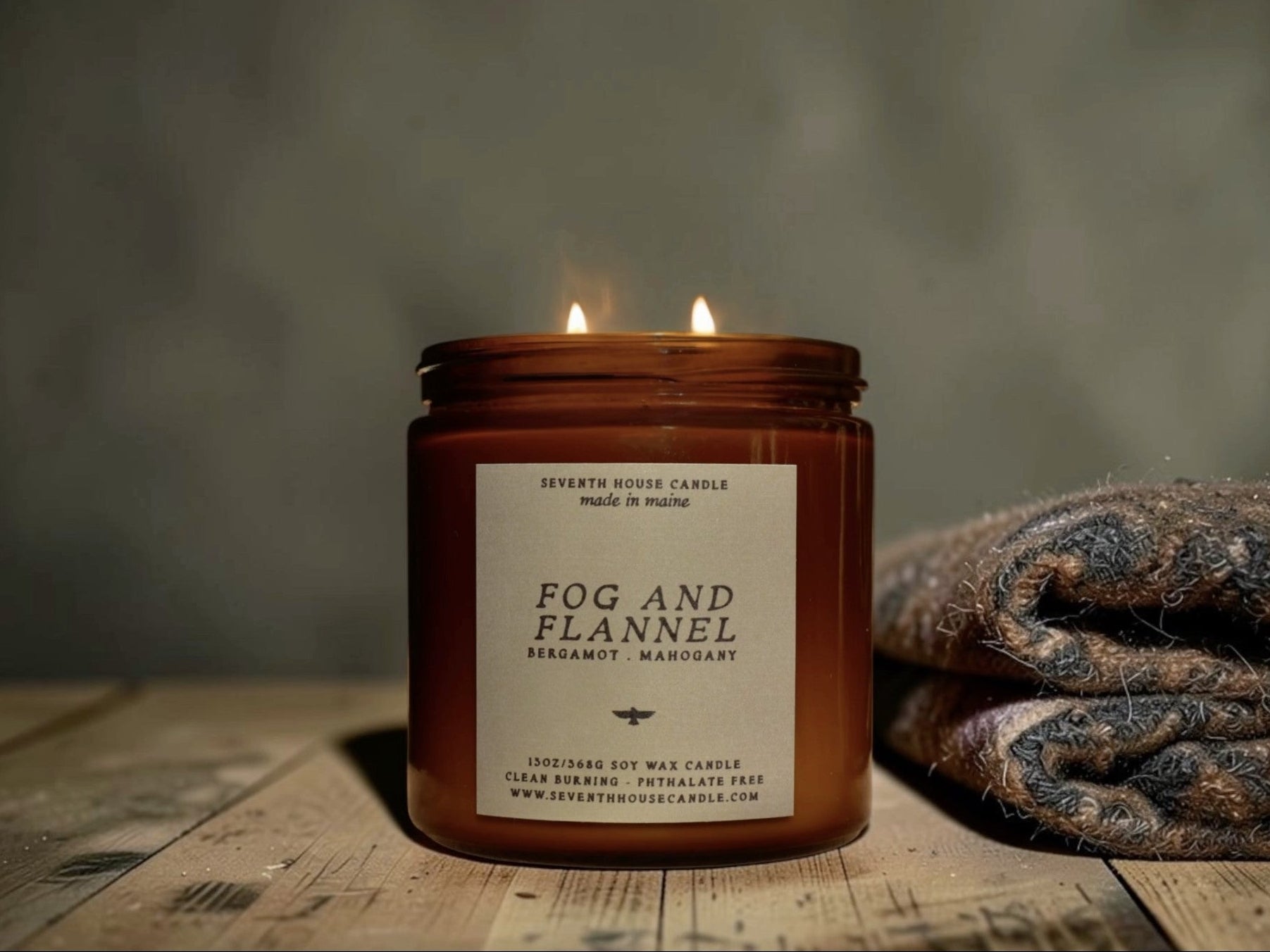 A lit amber glass jar candle labeled 'FOG AND FLANNEL' by Seventh House Candle, placed on a rustic wooden surface with a folded knit blanket beside it against a soft gray background.