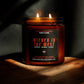 A lit amber glass jar candle with two flames, featuring a black label with red and white text that reads 'Wicked in the West' and 'Maple, Acorn, Clove'. The candle is placed on a wooden surface in dramatic, warm sunlight against a dark background.