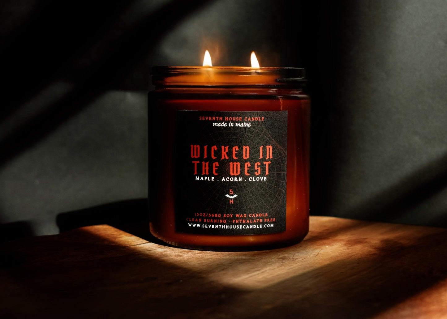 A lit amber glass jar candle with two flames, featuring a black label with red and white text that reads 'Wicked in the West' and 'Maple, Acorn, Clove'. The candle is placed on a wooden surface in dramatic, warm sunlight against a dark background.