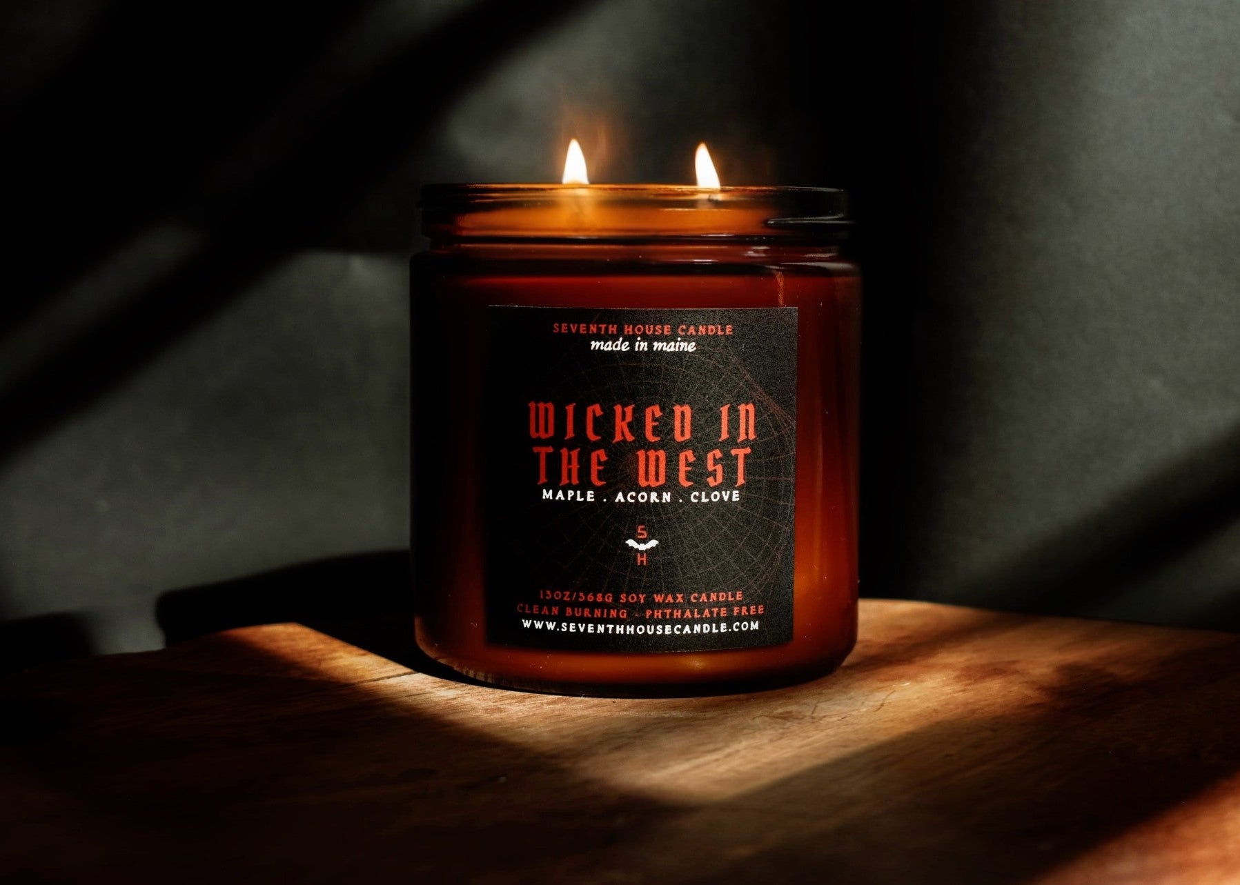 A lit amber glass jar candle with two flames, featuring a black label with red and white text that reads 'Wicked in the West' and 'Maple, Acorn, Clove'. The candle is placed on a wooden surface in dramatic, warm sunlight against a dark background.