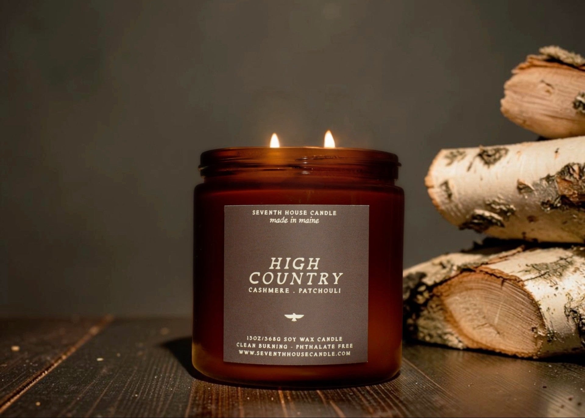 A lit amber glass jar candle labeled 'High Country' sits on a dark wooden surface beside stacked birch logs against a dark gray background.